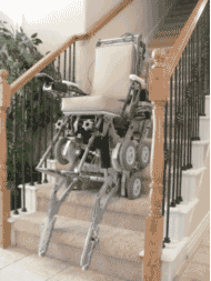 StairMaster on Stairs” shows an empty wheelchair resting on the second and third step of a carpeted home stairway.  A three spoke spider wheel with small diameter minor wheels at the end of the spoke is visible on the side of the wheelchair.  The wheelchair is balanced by a pair of skids on linkages at either side of the front of the wheelchair. The skids are resting on the edge of the first step. A rear caster and rear skids have been retracted, but are hidden behind the wheelchair. 